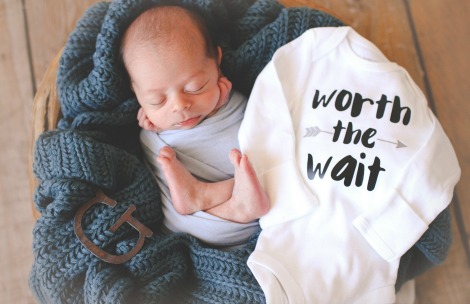 Sleeping newborn baby posed next to a onesie that reads 'Worth the Wait'
